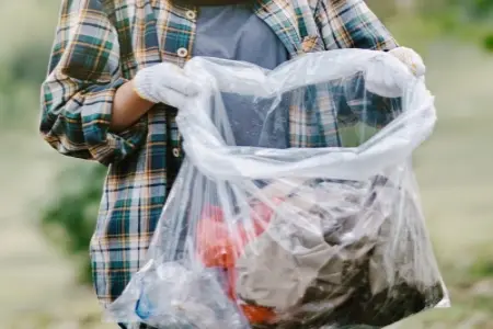 A person wearing a plaid shirt is holding a trash bag containing various types of trash. 