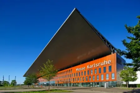An upward shot of the main entrance to the Karlsruhe Trade Fair Center. Green trees are in the foreground, and a blue sky shines in the background.