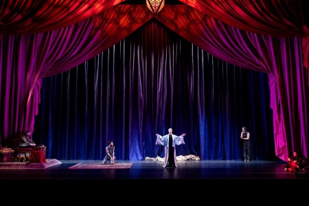 Eine Theateraufführung mit einem orientalischen Thema. Auf der Bühne sind zwei Schauspieler in farbenfrohen Kostümen zu sehen. Im Vordergrund steht ein Schauspieler, der in eine dramatische Pose eingefroren ist, während ein andere Darsteller im Hintergrund eine Szene spielt. Die Bühne ist mit detaillierten Kulissen und Requisiten dekoriert, die an 