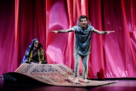 Eine Theateraufführung mit einem orientalischen Thema. Auf der Bühne sind zwei Schauspieler in farbenfrohen Kostümen zu sehen. Im Vordergrund steht ein Schauspieler, der in eine dramatische Pose eingefroren ist, während ein andere Darsteller im Hintergrund eine Szene spielt. Die Bühne ist mit detaillierten Kulissen und Requisiten dekoriert, die an 