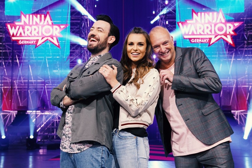 Die Moderatoren Frank Buschmann, Jan Köppen und Laura Wontorra vor den Ninja Warrior Bannern 